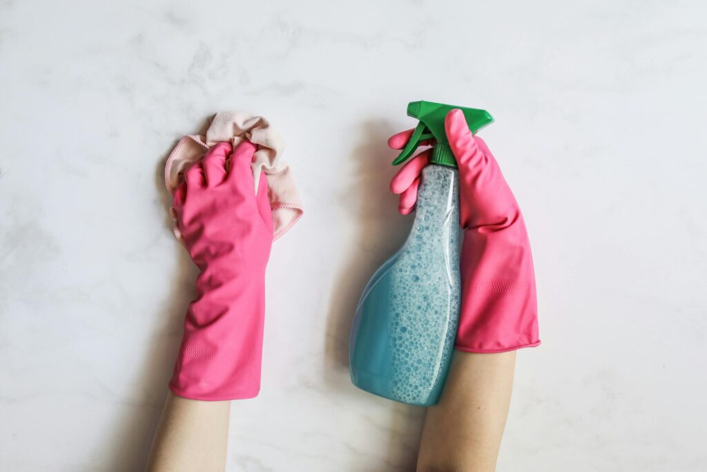 A pair of hands in pink gloves are cleaning with a spray bottle on a white background.